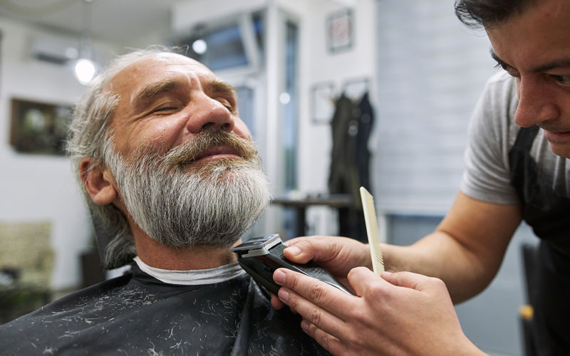 Barber trims smiling mans beard at a salon with a trimmer and comb.