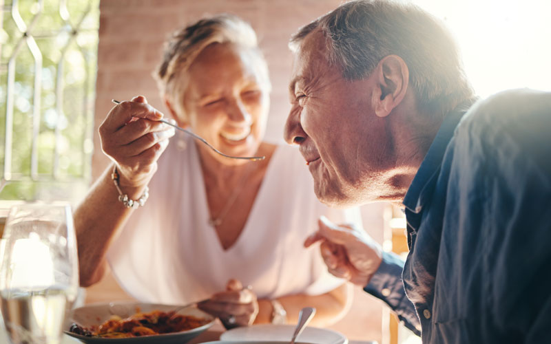 Elderly woman smiling and feeding a cheerful elderly man at an outdoor dining table.