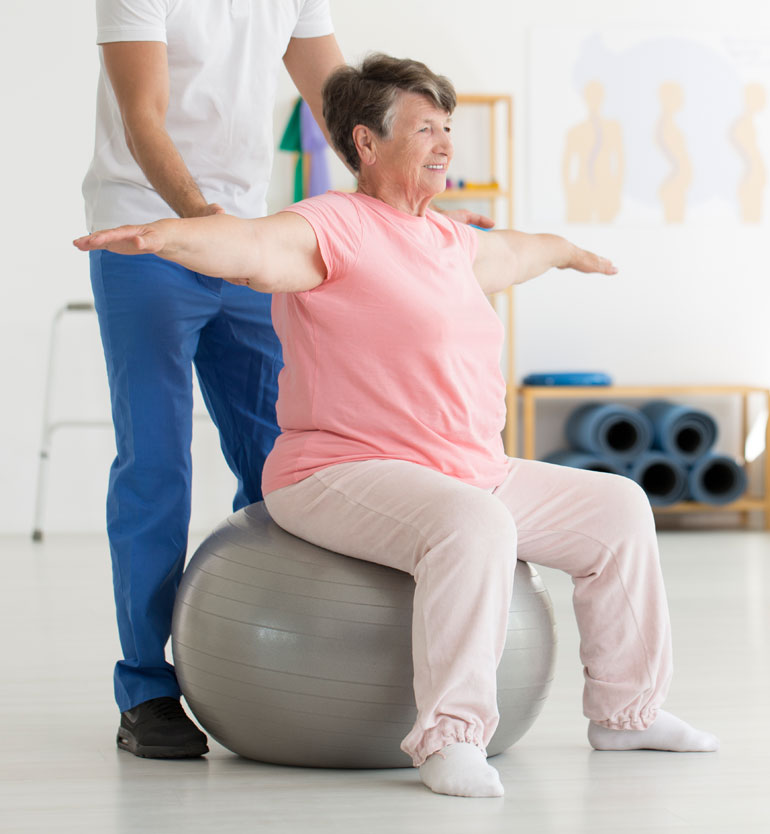 Elderly woman in pink shirt exercising on stability ball with support from therapist in blue pants.