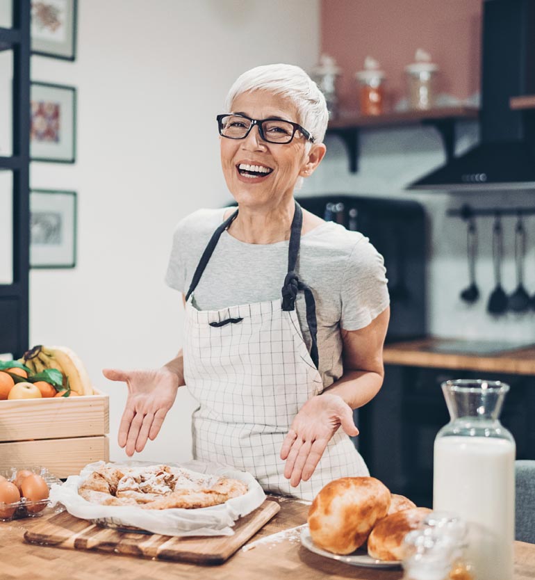 An elderly woman with grey hair and glasses smiling proudly at her freshly baked pastries in the kitchen.