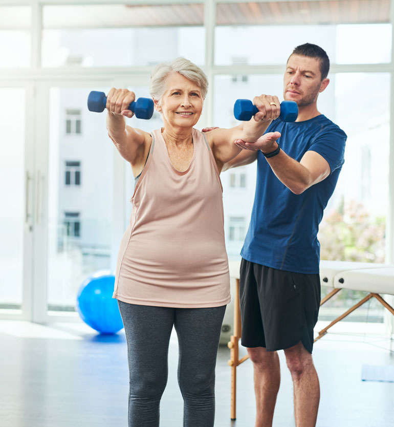 Elderly woman lifting blue dumbbells while being assisted by an instructor in a bright gym.