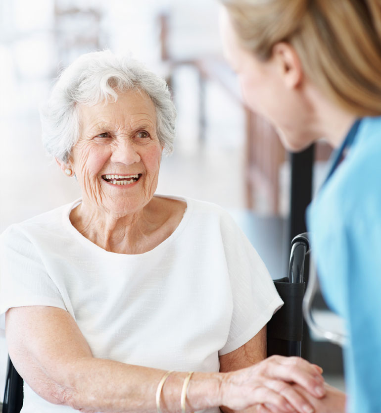 Elderly woman in a white shirt smiling warmly at a caregiver in blue scrubs during a visit