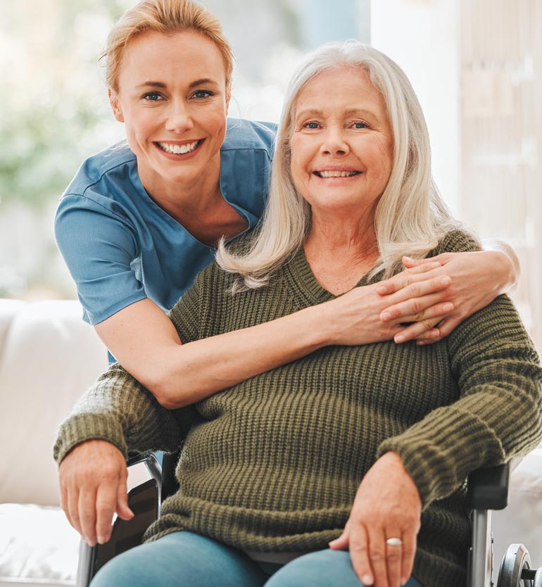 A cheerful caregiver hugging a smiling senior woman in a wheelchair, showcasing compassionate care.