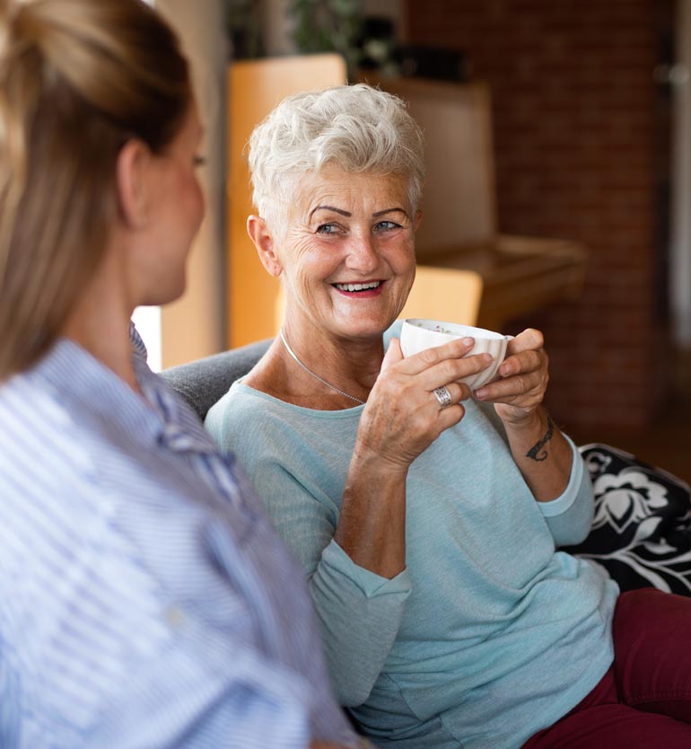 Senior woman joyfully talking with younger team member while holding a cup of tea indoors.