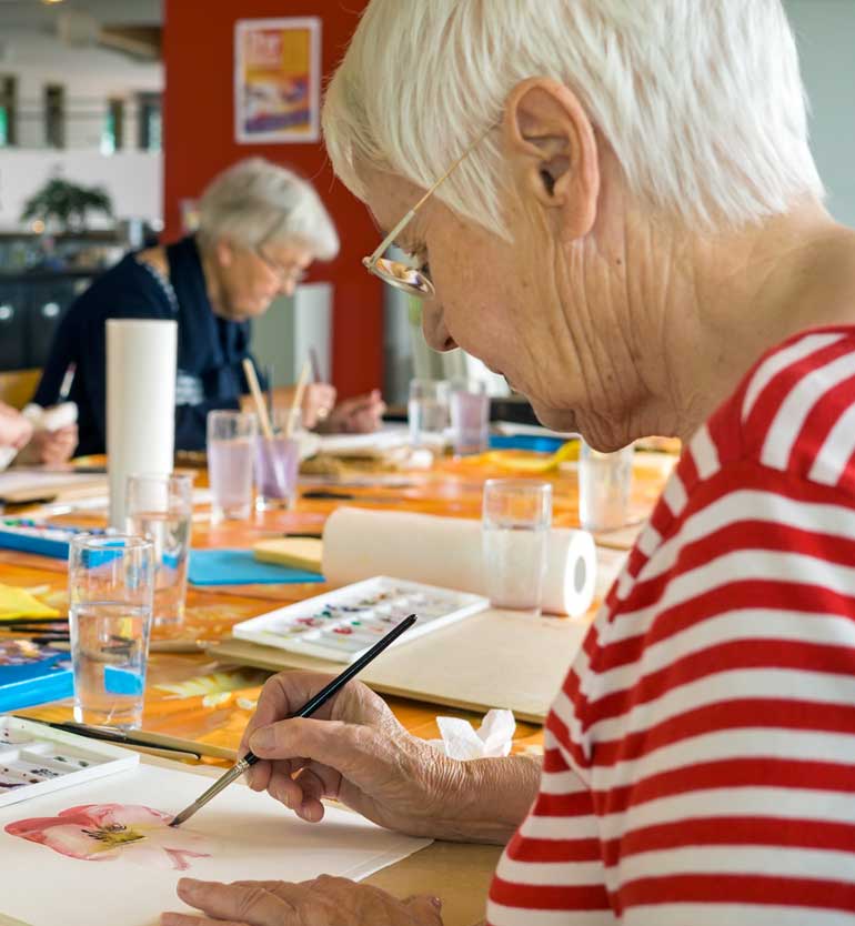 Senior woman painting watercolor at a table with other seniors in an art class setting.