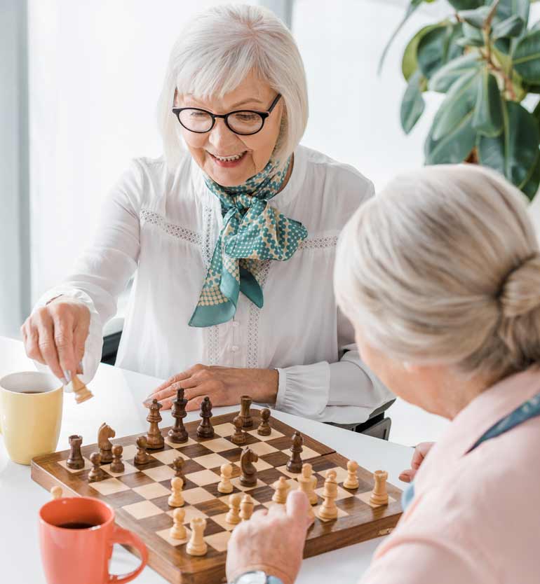 Two senior women happily engaged in a chess game at a bright indoor setting with coffee mugs nearby.