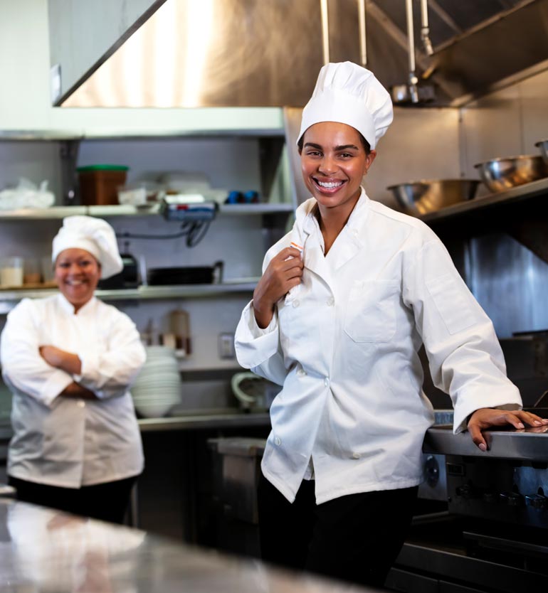 Two smiling chefs in white uniforms and hats working in a commercial kitchen with stainless steel equipment.